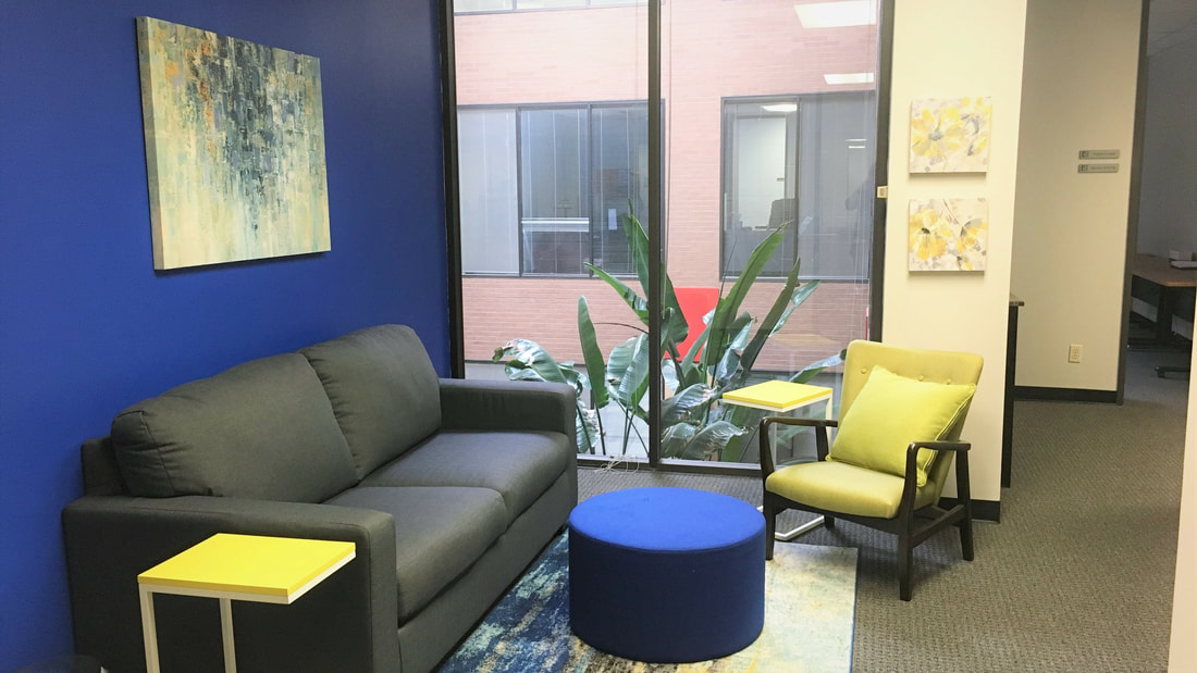 Open Client Lounge furnished with a couch, a chair with a note taking arm-rest, a round ottoman, and a side table. 