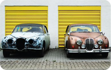 Two classic cars parked in front of yellow garage doors.