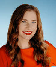 Head shot portrait of Sibbie Priestly smiling. She has red hair and is wearing an orange blouse.